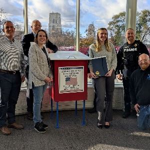 American Flag Retirement Drop Box displayed indoors with six people standing behind it, holding docu
